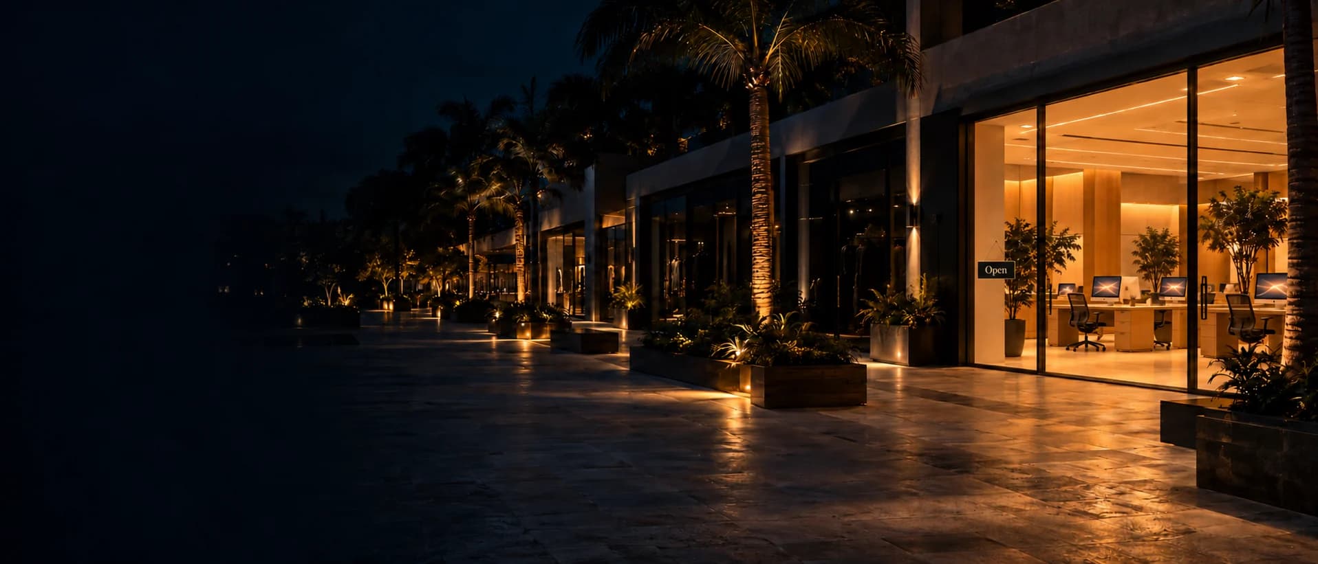 An upscale promenade at night, with palm trees and lit gardens: the shop windows are dark and only one office is lit, computers on and an Open sign on the door.