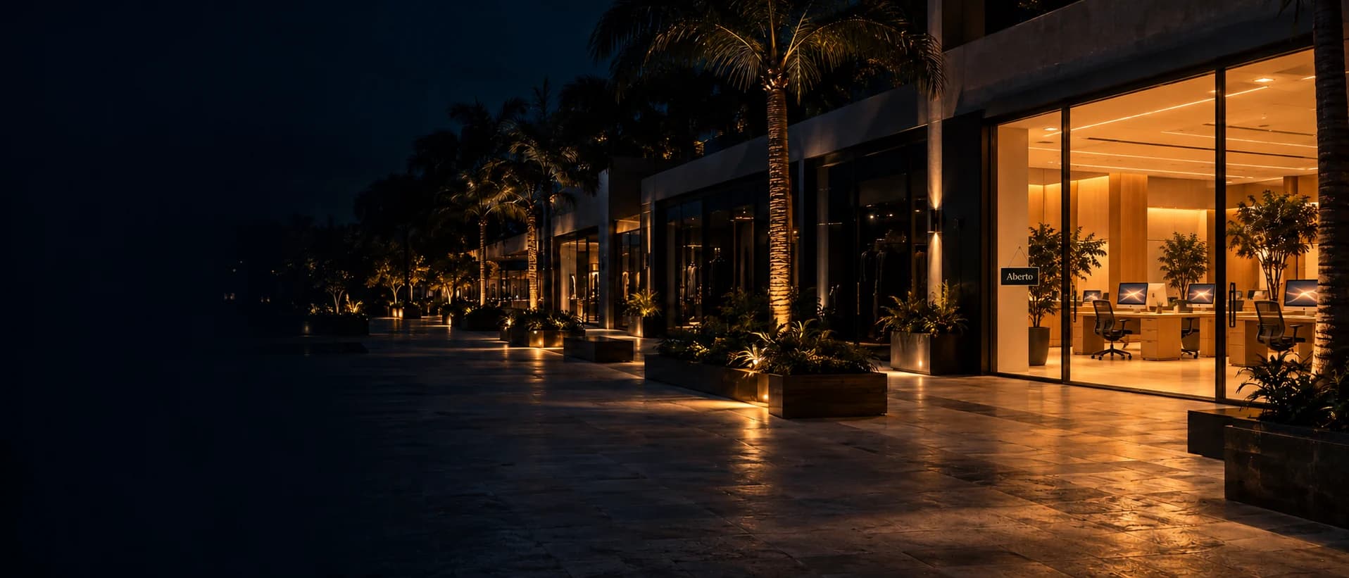 Calçadão de alto padrão à noite, com palmeiras e jardins iluminados: as vitrines escuras e só um escritório aceso, com os computadores ligados e a placa Aberto na porta.